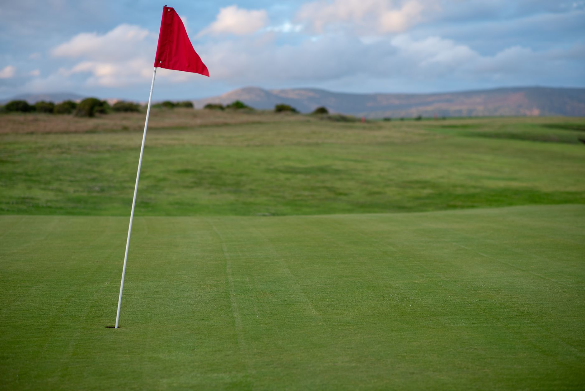 Drapeau rouge sur un mât blanc planté dans un green de golf, avec un parcours herbeux et des collines au loin sous un ciel nuageux.