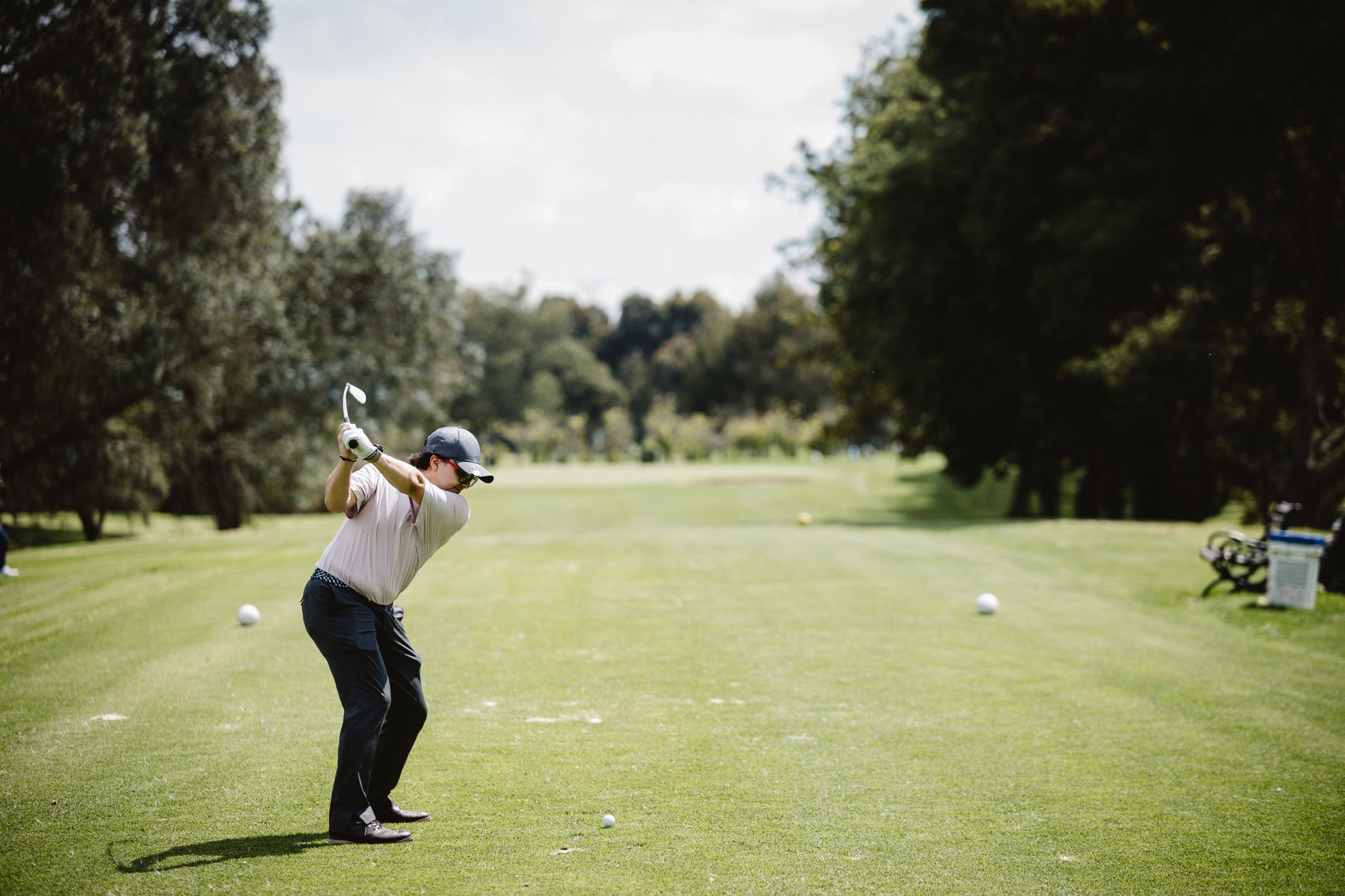Golfeur en polo clair et casquette au sommet de son élan sur un parcours de golf bordé d’arbres, avec des balles sur l’herbe.