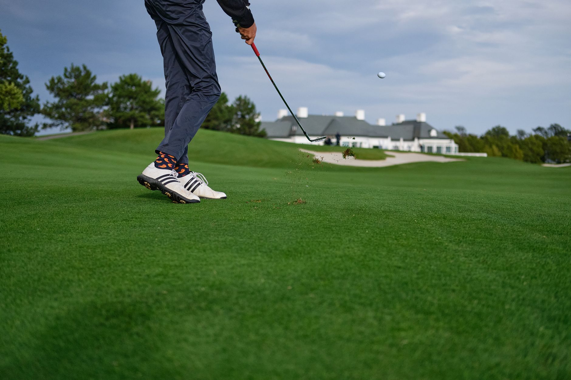 Golfeur vu de bas en train de frapper une balle avec un fer sur un fairway, un bunker et un bâtiment blanc en arrière-plan.