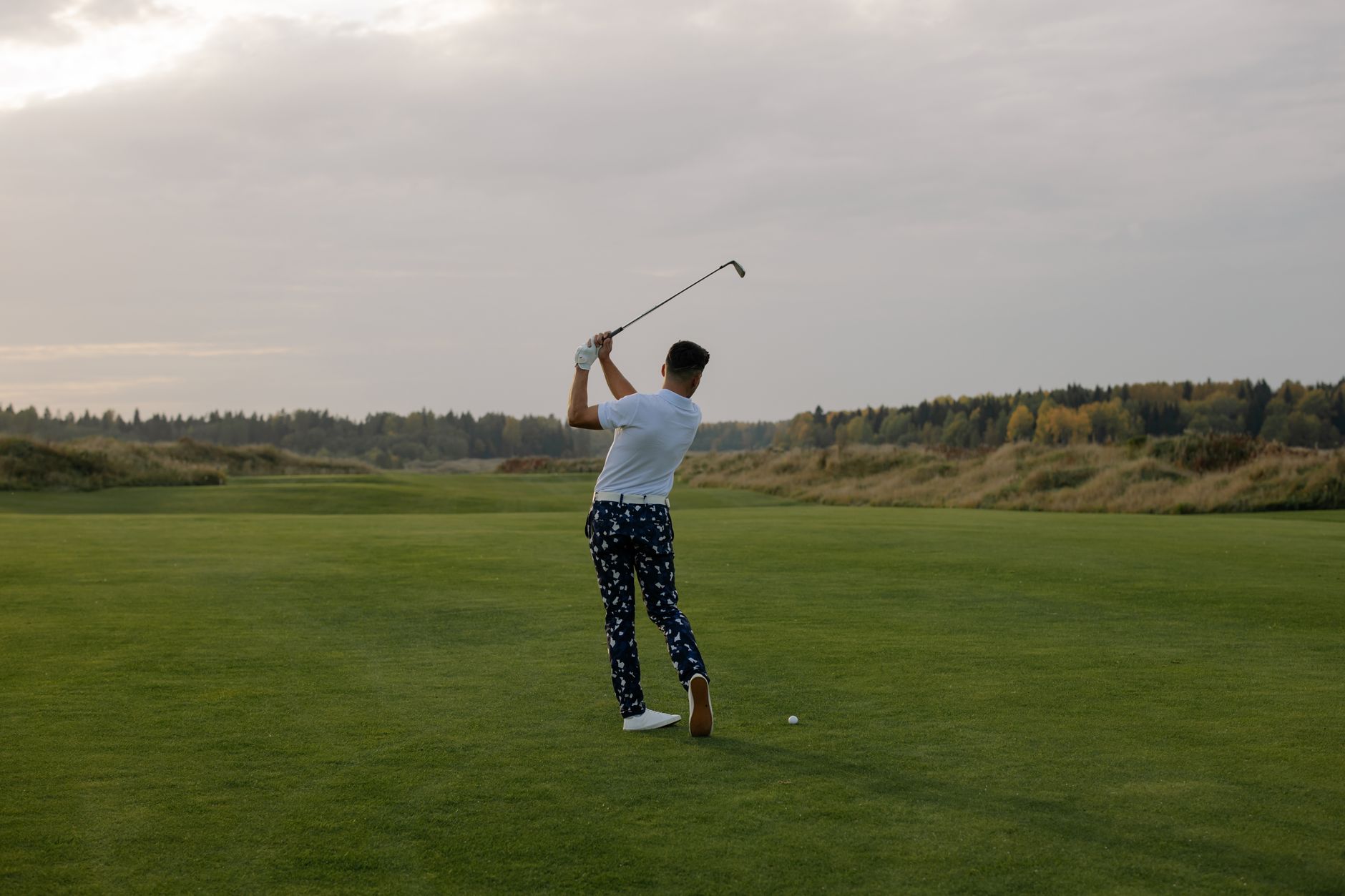 Golfeur vu de dos finissant son swing sur un fairway, avec des arbres aux teintes d’automne en arrière-plan.